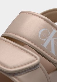 Κοντινή λήψη από μπεζ λουριά σανδαλιών με επενδεδυμένες άκρες και ορατές ραφές, με εν μέρει ορατό λογότυπο Calvin Klein σε λευκό χρώμα.