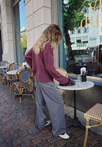 Bordeauxfarbener Strickpullover, weite graue Hosen und cremefarbene Schuhe. Marmor Tisch mit Besteck, umgeben von Korbstühlen und Kopfsteinpflaster.