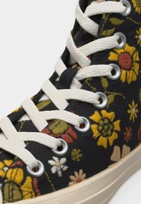 Baskets montantes en toile avec lacets blancs et œillets métalliques, ornées de motifs floraux brodés jaunes, orange et blancs sur tissu noir.