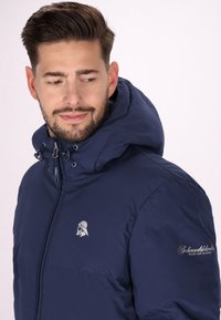 Marinblå puffjacka med huva, som har en silverfärgad logotyp på bröstet och en mönstrad detalj på ärmen. Självslät, lätt textur.