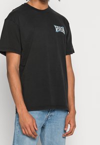 Camiseta de algodón negra de manga corta, con un logo en azul y blanco de "Tommy" en el lado izquierdo del pecho en un diseño rectangular.