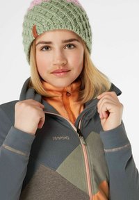 Beanie verde a maglia con accenti rosa; giacca in fleece grigia con design a blocchi di colore e strato interno arancione; dettagli con zip e marchio visibili.