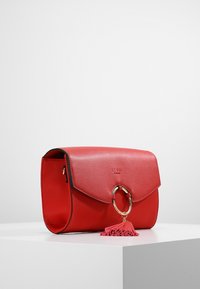 Borsa a tracolla rossa in pelle con superficie texturizzata, ferramenta in metallo dorato e un accento di nappina rosa sul lato. Presenta una chiusura a patta.