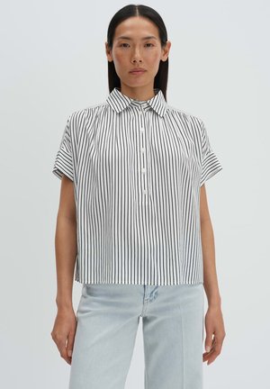 Femme portant une chemise blanche à manches courtes à boutons avec des rayures noires verticales et un jean bleu clair, debout devant un fond uni.