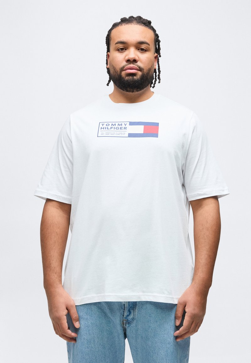 Άνδρας με πλεξούδες, φορώντας άσπρο T-shirt Tommy Hilfiger και ανοιχτό μπλε τζιν, στέκεται μπροστά σε απλό ανοιχτό φόντο.