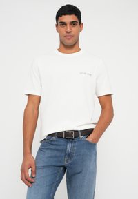 Man draagt een wit T-shirt met korte mouwen met kleine tekst en een blauwe spijkerbroek, staat met één hand in de zak tegen een effen achtergrond.