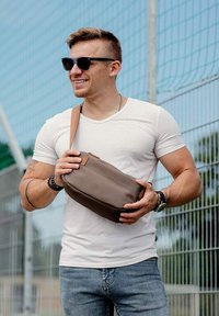 Hnědá kožená crossbody taška s hladkým povrchem, obdélníkovým tvarem a popruhem; drží ji osoba v bílem tričku a džínech venku.