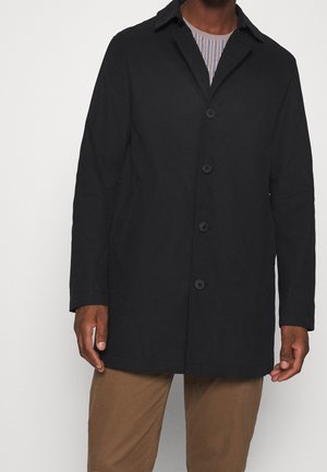 Homme portant un manteau noir à boutons par-dessus une chemise rayée et un pantalon marron, debout devant un fond clair uni.