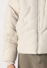 Beige puffjacka med kviltade stjärnmönster, ribbade ärmslut och dragkedja fram. Mjukt tyg med en slät yta.