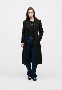Cappotto nero lungo con design strutturato, indossato sopra una t-shirt nera e jeans a gamba larga blu, abbinato a scarpe nere.