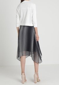 Cardigan blanco con mangas tres cuartos sobre un vestido de lunares en blanco y negro. El vestido presenta un dobladillo asimétrico y una textura transparente.
