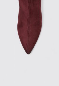 Zapato de ante burdeos con punta afilada, costuras a lo largo de los laterales y textura suave, que muestra una silueta elegante y alargada.