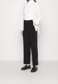 Witte, lange mouwen shirt over een geribbelde witte top, gecombineerd met zwarte wijde broek met glanzende zijaccenten. Zwarte loafers maken de look af.