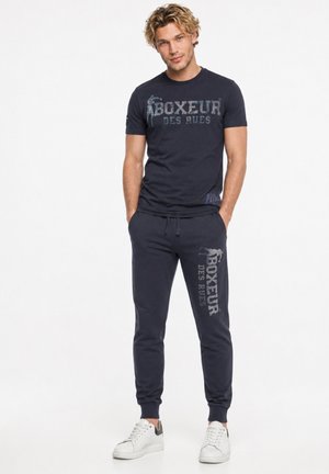 Junger Mann mit den Händen in den Taschen, der ein dunkelblaues "Boxeur des Rues" T-Shirt und passende Jogginghosen mit weißen Sneakern vor weißem Hintergrund trägt.