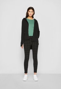 Svart hettegenser med glidelås, grønn T-skjorte, svarte leggings med rutemønster, og hvite sneakers. Glatt stoff, tettsittende design og kontrasterende farger.