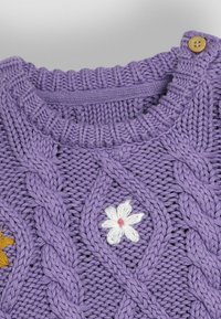Lila gestrickter Pullover mit einem Zopfmuster, weißer Blumenstickerei mit pinkem Mittelpunkt und einem Holzknopf an der Schulter.