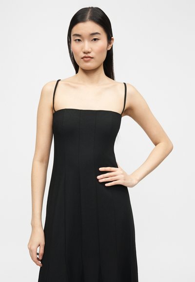 Jeune femme aux longs cheveux noirs portant une robe noire sans manches à fines bretelles, posant avec une main sur la hanche sur un fond uni.