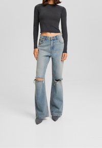 High-waisted, light blue denim jeans with large knee rips, flared legs, and a raw hem, paired with a dark cropped long-sleeve top.