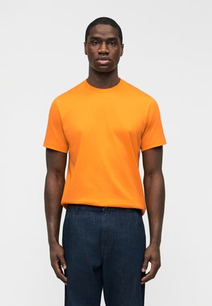 Egy férfi, aki egyszerű, élénk narancssárga rövid ujjú pólót és sötétkék nadrágot visel, egy egyszerű világosszürke háttér előtt áll.