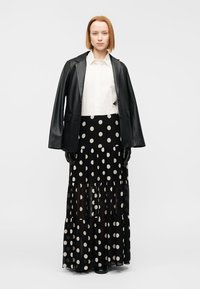 Μαύρο δερμάτινο μπλέιζερ πάνω από ένα λευκό πουκάμισο, συνδυασμένο με μαύρη μακριά φούστα με πουά, διακοσμημένη με διαφανή στρώματα και βολάν. Μαύρα γάντια.