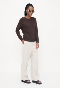Pull marron en tricot doux, associé à un pantalon large beige clair. Le mannequin porte des lunettes de soleil claires et des chaussures noires épaisses avec des accents violets.