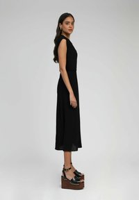 Robe plissée noire à manches courtes, longueur genou. Portée avec des sandales compensées noires épaisses avec un aspect bois et des accents métalliques.