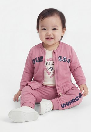 Tout-petit assis et souriant, portant une veste et un pantalon roses Guess avec des chaussures blanches sur fond blanc.