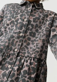 Haljina košulja s ovratnikom u sivom i ružičastom uzorku leoparda. Ima gumbe sprijeda i dizajn sa skupljenim strukom za dodatnu teksturu.