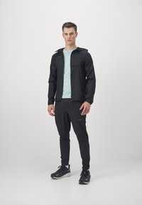 Veste légère noire avec capuche, portée sur une chemise vert menthe, associée à un pantalon noir et des chaussures de course. Texture lisse et design épuré.