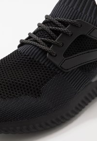 Sneaker sportivo nero con tomaia in rete, motivo testurizzato, lacci neri e suola in gomma spessa con design a righe.