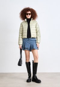 Sieviete ar cirtainiem matiem un saulesbrillēm, ģērbusies bēšā pūkainā jakā, melnā augstajā t-kreklā, džinsā ar puķēm, melnās zeķubiksēs, turot melnu somu.