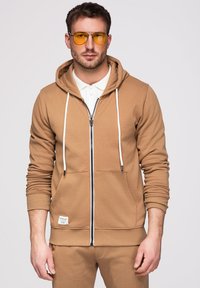 Homme portant un sweat à capuche beige zippé et un pantalon assorti, des lunettes de soleil teintées jaunes et une chemise blanche à col en-dessous.
