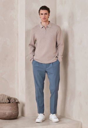 Ein junger Mann steht drinnen, trägt einen beigen Pullover mit Reißverschlusskragen, blaue maßgeschneiderte Hose und weiße Sneakers, die Hände in den Taschen neben einem Korb mit Strickwaren.