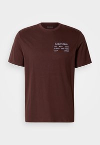 Camiseta de algodón marrón con mangas cortas, cuello redondo y texto impreso en blanco y azul claro. El texto incluye detalles de la marca y la dirección.