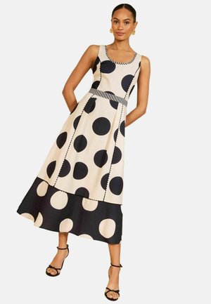Femeie purtând o rochie crem fără mâneci cu buline mari negre și tiv negru cu buline crem, asortată cu sandale negre cu barete.