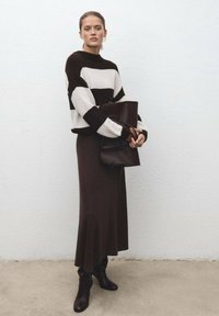 Suéter de punto a rayas en marrón y crema, combinado con una falda midi acampanada de color marrón oscuro. La modelo sostiene un bolso de mano marrón y lleva botas altas.