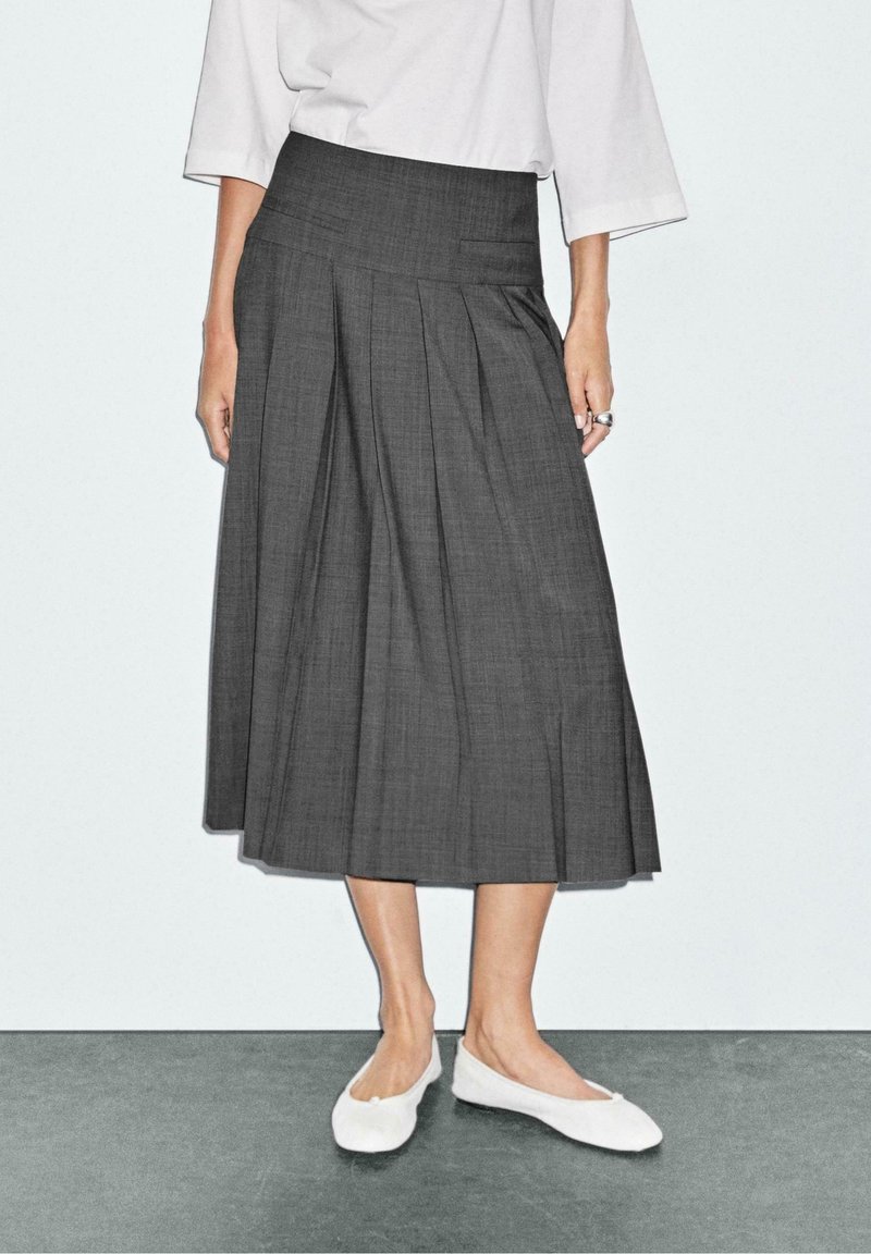 Femme portant une jupe midi plissée gris foncé taille haute avec des ballerines blanches, debout contre un mur blanc uni.