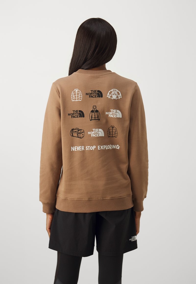 Personne portant un sweat-shirt marron avec des icônes de matériel de plein air et le texte « Never Stop Exploring », associé à un short noir et des leggings.