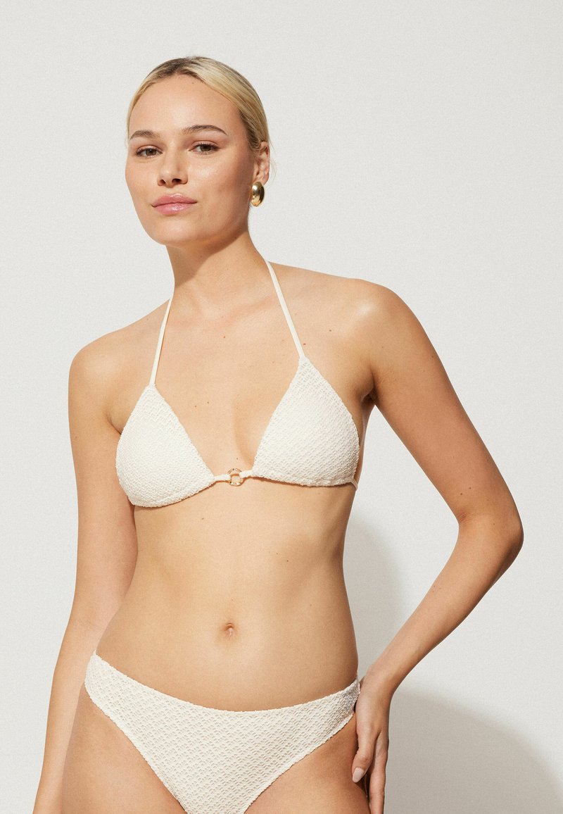 Bílé texturované bikini s trojúhelníkovým topem a zlatým kroužkem uprostřed, doplněné odpovídajícím spodním dílem; minimalistický design.