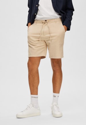 Homme portant un short beige à cordon, des chaussettes blanches, des baskets blanches, un t-shirt blanc et une chemise ouverte bleu marine les mains dans les poches.