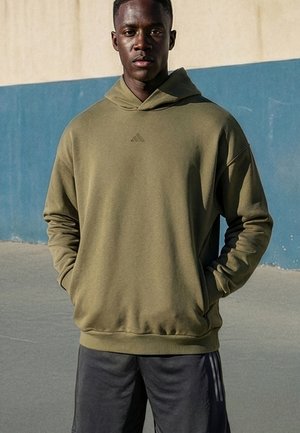 Junger Mann mit einem olivgrünen Adidas-Hoodie und schwarzen Adidas-Hosen steht im Freien vor einer beige-blauen Wand.