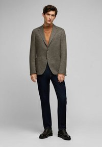 Grau melierter Blazer mit Reverskragen, zwei vorderen Taschen und einem Einzelknopfverschluss, kombiniert mit einem braunen Rollkragenpullover und dunklen Jeans.