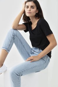 Czarny t-shirt krótki rękaw w połączeniu z jasnoniebieskimi spodniami o wysokim stanie. Model siedzi z jedną nogą zgiętą, nosząc białe trampki. Gładka faktura materiału.