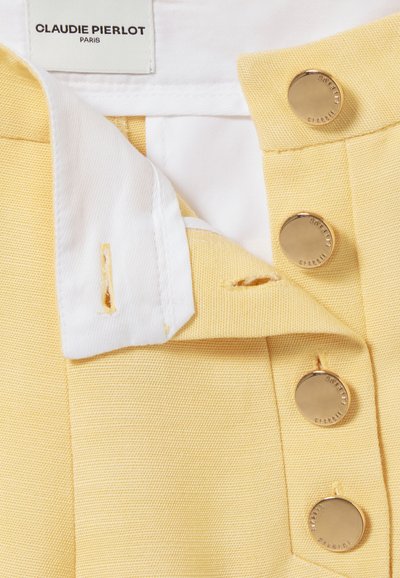 Tissu jaune avec une finition texturée, arborant un col blanc et trois boutons dorés. L'étiquette indique "CLAUDIE PIERLOT PARIS."