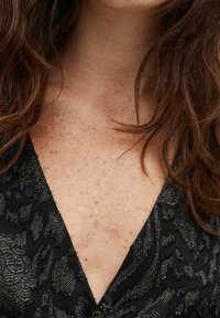 Zwarte jurk met een diepe V-hals en textiel met een bloemenmotief. Haar is golvend en van gemiddelde lengte, waardoor de halslijn wordt omlijst.
