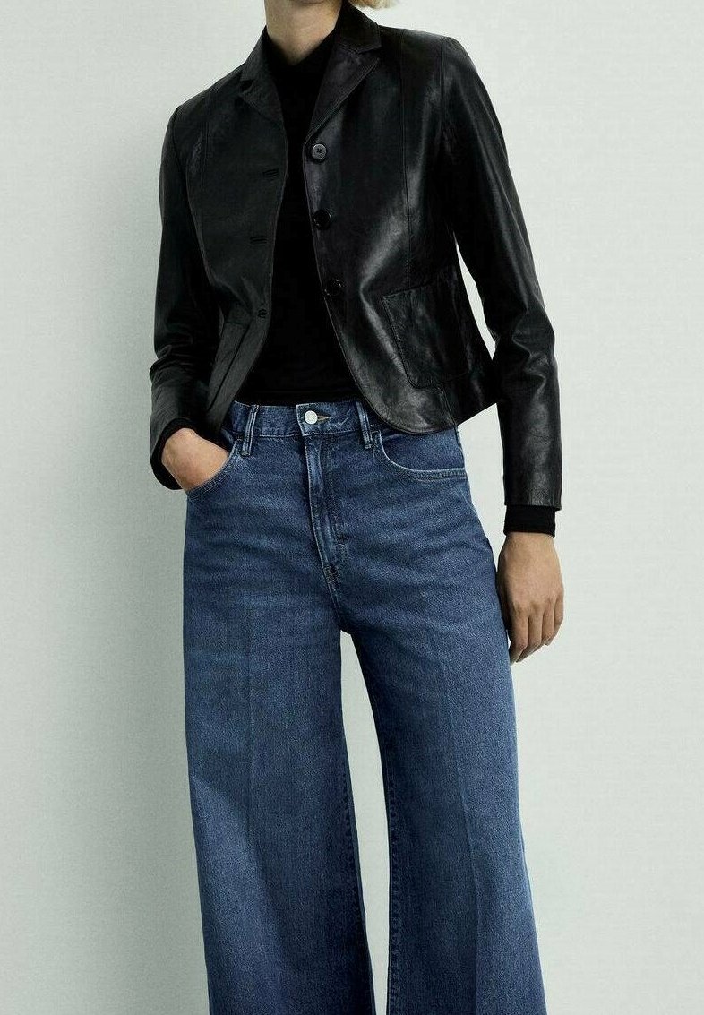 Personne portant un blazer en cuir noir sur un haut noir et un jean bleu taille haute à jambes larges, debout avec une main dans la poche devant un fond uni.