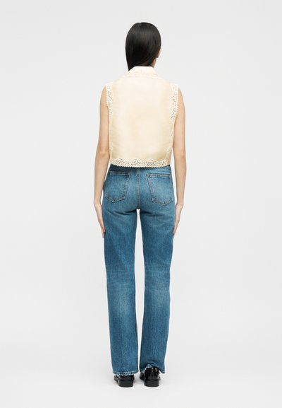 Γυναίκα με μακριά σκούρα μαλλιά που φοράει αμάνικη μπεζ μπλούζα με δαντέλα και ψηλόμεσα κανονικά μπλε τζιν, στεκόμενη με την πλάτη προς τα εμπρός.