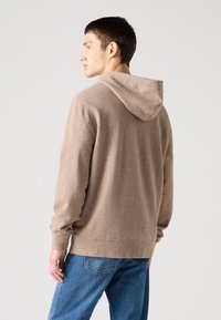 Felpa marrone con un aspetto tessurizzato, dotata di cappuccio e maniche lunghe, abbinata a jeans blu. Il materiale sembra morbido e informale.