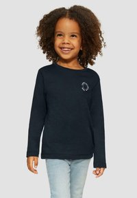 Glunderend kind met krullend haar, gekleed in een zwart langemouwen shirt en lichtblauwe jeans, staand voor een effen achtergrond.