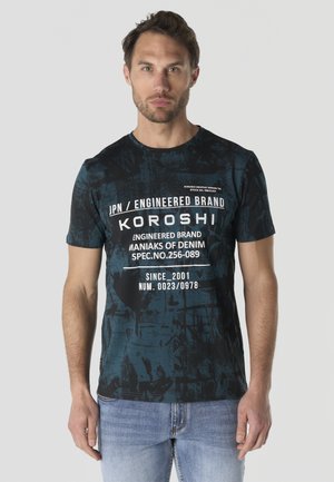 Hombre con una camiseta gráfica negra y azul con texto blanco y vaqueros azul claro, de pie frente a un fondo gris liso.
